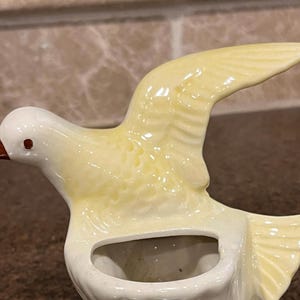 Könnte beinhalten: Keramik-Vogelfigur im Flug, mit cremefarbenem Kopf und Körper und einem gelben Flügel. Der Vogel hat einen braunen Schnabel und ein kleines, rotes Auge. Die Figur hat einen ausgehöhlten Bereich, möglicherweise zum Halten kleiner Gegenstände.