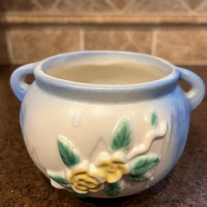 May include: A small, round, ceramic bowl with two handles. The bowl is primarily white with a light blue rim and handles. It features a floral design with yellow flowers and green leaves.