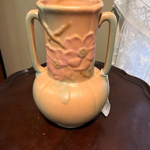 May include: Peach-colored ceramic vase with two handles and a bulbous base. The vase features a floral design in pink and a scalloped rim. The handles and the rim have a light green hue.