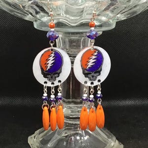 May include: A pair of dangle earrings featuring a white circular charm with a Grateful Dead Steal Your Face logo in orange, purple, and white. The charms are attached to silver chains with orange, white, and blue beads.