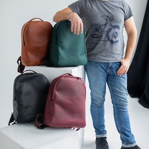 May include: Four leather backpacks in different colours: brown, green, black, and burgundy. The backpacks are displayed on a white cube, with a person leaning against them. The person is wearing a grey t-shirt and blue jeans.