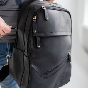May include: Black leather backpack with gold-coloured zips and details. The backpack features multiple compartments and a top handle. The bag is held by a person wearing a grey t-shirt and blue jeans.