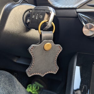 May include: A black leather key chain with a brass ring and a brass button. The keychain is attached to a car key.