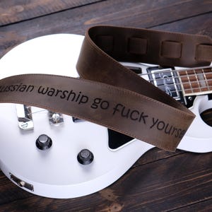Può includere: Un basso elettrico bianco con un cinturino in pelle marrone. Il cinturino riporta la scritta "Russian warship go fuck yourself". La chitarra è su una superficie di legno scuro.