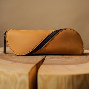 Può includere: Un astuccio per occhiali in pelle color cuoio con chiusura a cerniera nera. L'astuccio ha un design curvo ed è cucito con filo nero. È appoggiato su una superficie di legno marrone chiaro.