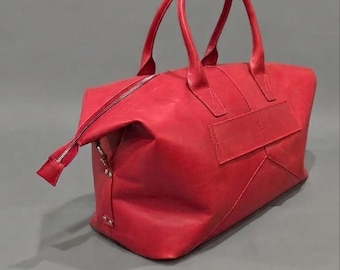 May include: A bright red leather duffel bag featuring two top handles, a silver-toned metal zip closure, and a rectangular patch detail on the side. The bag has a structured base with visible stitching and metal rivets along the side panels.