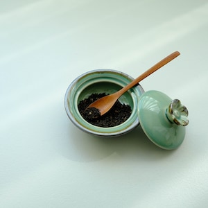 May include: A green ceramic tea jar with a flower-shaped lid and a wooden spoon inside. The jar is filled with loose leaf black tea.