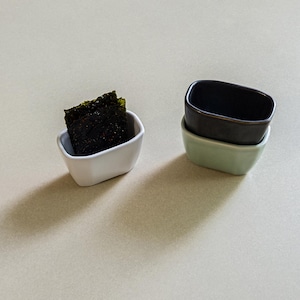 以下が含まれることがあります： 白い陶器のボウルに入った長方形の濃い緑色の乾燥海苔と、マットブラックと淡いセージグリーンの2つの小さな陶器のボウルが積み重なったセット。
