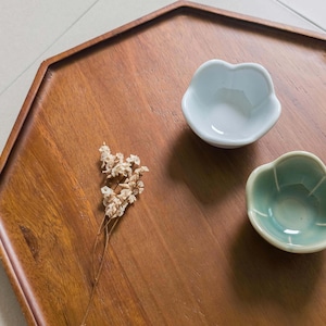 May include: A wooden serving tray with a hexagonal shape, holding two small, flower-shaped ceramic bowls in light blue and green. A sprig of dried flowers rests on the tray. The tray is made of a warm-toned wood.