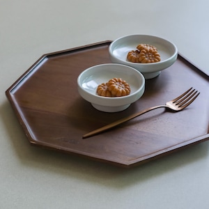 May include: A dark brown, octagonal wooden tray holds two small white bowls with baked goods and a bronze fork. The bowls are on pedestals. The baked goods are a golden brown color.