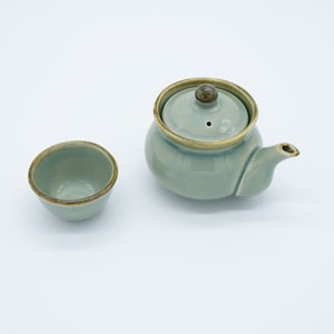 May include: A small, light green ceramic teapot with a lid and a matching teacup. The teapot has a brown rim and a small brown knob on the lid.