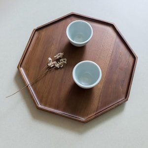 Könnte beinhalten: Ein achteckiges Serviertablett aus Holz mit dunkelbraunem Finish. Zwei kleine, hellblaue Keramikbecher stehen auf dem Tablett, zusammen mit einem Zweig getrockneter Blumen. Das Tablett steht vor einem neutralen Hintergrund.