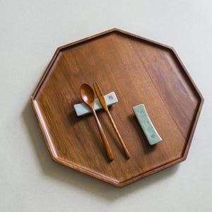Könnte beinhalten: Ein hölzernes Serviertablett mit achteckiger Form. Ein Holzlöffel und eine Gabel liegen auf dem Tablett, zusammen mit zwei Keramik-Essstäbchenhaltern. Das Tablett und die Utensilien sind warmbraun.