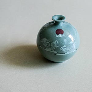 Może przedstawiać: Okrągły, jasnoniebieski wazon ceramiczny z wykończeniem typu spękana glazura. Wazon ozdobiony jest białym wzorem graficznym przedstawiającym dwa lecące żurawie oraz małym, ciemnoczerwonym motywem słońca w pobliżu wąskiego, okrągłego otworu.