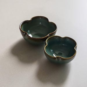 以下が含まれることがあります： 独特な四つ葉の形をした、2つのティール色のセラミックボウル。ボウルは光沢のある仕上げで、濃い茶色の縁があります。ボウルはサイズが異なり、明るい色の表面に置かれています。
