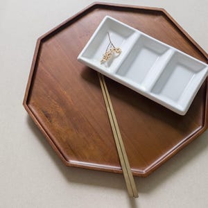 Peut inclure: Un plateau de service octogonal en bois avec un plat blanc divisé et une paire de baguettes. Le plateau est de couleur marron foncé et le plat comporte trois compartiments. Une petite brindille de fleurs séchées repose dans le plat.