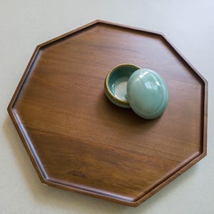 May include: A wooden, octagonal tray with a dark brown finish. A small, light green ceramic container with a lid sits on the tray. The container has a gold rim and a white interior. The lid is oval-shaped.