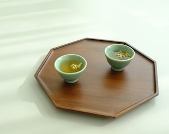 May include: Two small green teacups filled with light yellow tea and flower buds sit on a dark brown octagonal wooden tray.