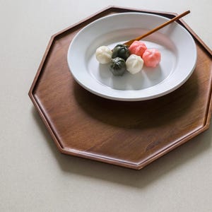 May include: A wooden, octagonal tray holds a white plate with colorful, round pastries and a wooden spoon. The pastries are white, pink, and green. The tray is a rich, dark brown color.