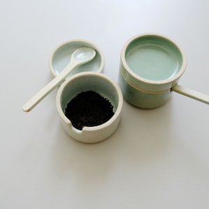 Könnte beinhalten: Zwei Keramikschalen mit weißem und hellgrünem Glase. Eine Schale enthält einen kleinen weißen Löffel und die andere Schale enthält eine kleine Menge schwarzer Teeblätter.