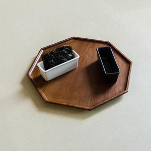 以下が含まれることがあります： 滑らかな茶色の木目仕上げの八角形の木製サービングトレイ。トレイの上には、濃い緑色の乾燥海苔が入った長方形の白いセラミック容器と、空の長方形の黒いセラミック容器が置かれています。