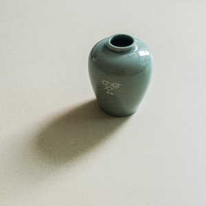 May include: A small, rounded ceramic vase in a soft sage green glaze with a subtle crackle finish. The vase features a delicate, hand-painted white floral motif on the front and a narrow, circular opening at the top with a smooth, rounded rim.
