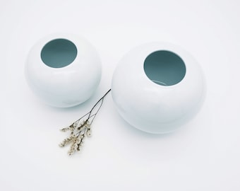 May include: Two white ceramic vases in the shape of spheres with a round opening at the top. The vases are sitting on a white surface.