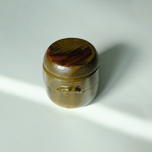 Peut inclure: Un petit pot rond en céramique verte avec un couvercle. Le couvercle est décoré de motifs marrons et orange.