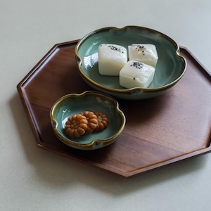 以下が含まれることがあります： 六角形の木製サービングトレイには、2つの小さなティール色のボウルが置かれています。1つのボウルには、黒いゴマをトッピングした3つの白い正方形の菓子が入っています。もう1つのボウルには、いくつかの小さくて丸い茶色のクッキーが入っています。トレイはニュートラルな背景に置かれています。