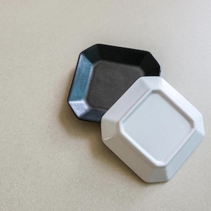 Könnte beinhalten: Zwei kleine, quadratische Keramikplatten mit abgeschrägten Kanten. Eine Platte ist schwarz, die andere weiß. Die weiße Platte ist umgedreht und zeigt ihre Unterseite. Die Platten dienen wahrscheinlich zum Servieren von Vorspeisen oder kleinen Portionen.