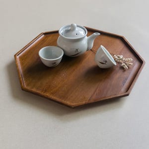 以下が含まれることがあります： 木製の八角形のサービングトレイには、蓋付きの小さな白いティーポット、お揃いのティーカップ2つ、ドライフラワーが置かれています。 ティーポットとカップには繊細な黒いデザインがあります。 トレイは暖かく、濃い茶色です。
