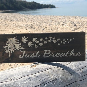 May include: A wooden sign with the words "Just Breathe" in white lettering. The sign features a carved marijuana leaf design with smaller leaves blowing in the wind.