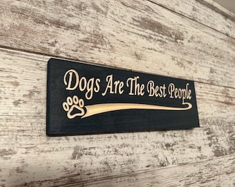 May include: A dark blue wooden sign with the text "Dogs Are The Best People" in white lettering. A paw print is carved into the wood above the text.