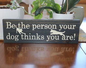 May include: A dark gray wooden sign with white text that reads "Be the person your dog thinks you are!" with two white silhouettes of dogs on either side of the text.