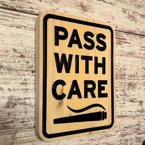 May include: A rectangular wooden sign with the words "PASS WITH CARE" in bold black letters. The sign has a black border and a black graphic of a lit cigarette. The background is a light wood grain.