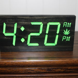 May include: A black wooden digital clock displaying "4:20" in bright green numerals. The clock also shows "AM" and "PM" with a cannabis leaf symbol. The clock is a rectangular shape and sits on a wooden surface.
