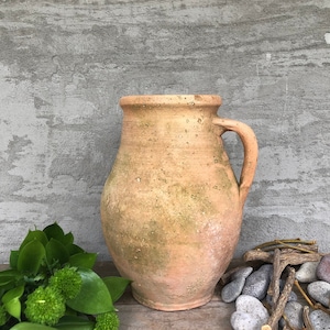 May include: A terracotta pitcher with a rustic, aged appearance. The pitcher has a wide, round body and a single handle. The pitcher is sitting on a wooden surface with green foliage and gray stones in the foreground.
