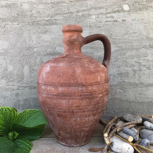 May include: A terracotta pitcher with a wide mouth and a handle. The pitcher has a rustic, hand-crafted look and is decorated with a series of horizontal lines.