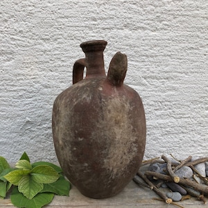 May include: A rustic brown clay jug with a handle and a spout. The jug is sitting on a wooden surface with green leaves and twigs.