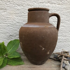May include: A brown earthenware jug with a handle. The jug has a rustic, aged appearance and is made of clay.