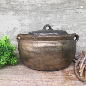 以下が含まれることがあります： ふたと2つの取っ手がついた、大きな茶色の土製の鍋。鍋は木製の表面の上に置かれています。