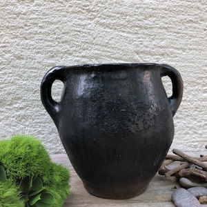 May include: A black ceramic pot with two handles. The pot has a rustic, aged look and is perfect for displaying plants or flowers.
