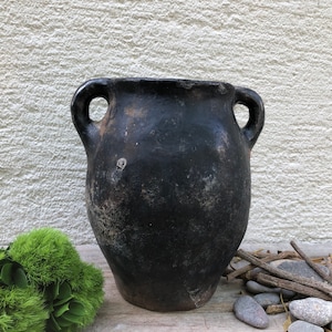 May include: A black ceramic pot with two handles. The pot has a rustic, aged look and is perfect for displaying plants or flowers.