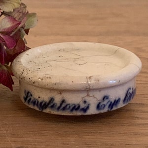 Può includere: Coperchio di barattolo in ceramica bianca con scritta blu che recita "Singleton's Eye Lotion".