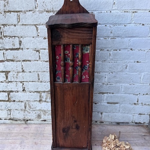 May include: A vintage wooden wall mounted storage cabinet with a red and white floral patterned fabric insert. The cabinet has a dark brown wood finish and a simple design.