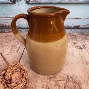 Puede incluir: Jarra de cerámica de dos tonos con asa y boquilla. La parte superior de la jarra es de color marrón oscuro, con un color beige claro en los dos tercios inferiores. El asa es de color beige y marrón.