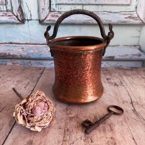 May include: A small, antique copper bucket with a dark handle. The bucket has a hammered texture and sits on a wooden surface next to a dried rose and a vintage key. The bucket is a decorative item.