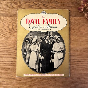 May include: A vintage book titled "The Royal Family Golden Album" with a black and white photo of the royal family on the cover. The book features a gold background and the text "143 Photographs" and "First Birthday Pictures of Prince Charles".
