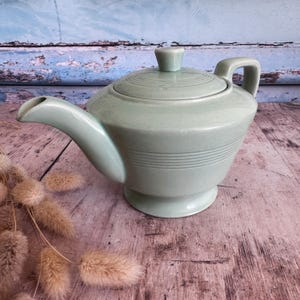 Könnte beinhalten: Eine hellgrüne Keramik-Teekanne mit Deckel und Henkel. Die Teekanne hat einen Ausguss und ist mit horizontalen Linien um den Körper verziert. Die Teekanne steht auf einer Holzoberfläche.