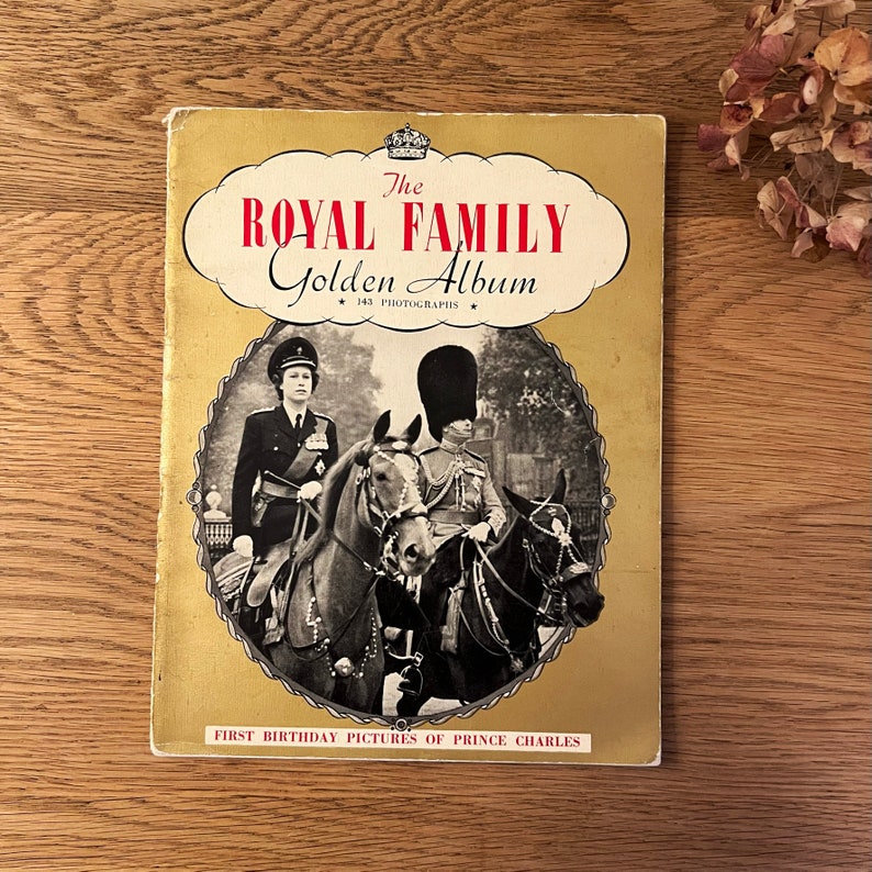 May include: A vintage book titled "The Royal Family Golden Album" with a gold cover and a black and white photo of Queen Elizabeth II and Prince Philip on horseback. The book features the text "143 Photographs" and "First Birthday Pictures of Prince Charles".